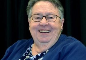 A headshot of an older woman smiling brightly. She has short grey and brown hair, rectangular tortoise glasses and is wearing a blue shirt.