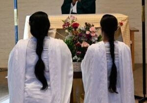 Two women with long dark braids and wearing long white garments kneel before a flower-bedecked altar. A priest is partially visible standing behind the altar.