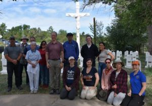 Fourteen men and women pose in front of the Loretto Heights Cemetery, with the large white crucifix and gravestones visible in the background.