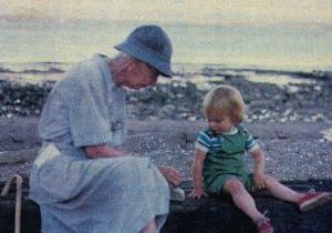 An older woman, with a cane propped behind her, stacks pebbles on the log on which she sits, while the toddler next to her watches.