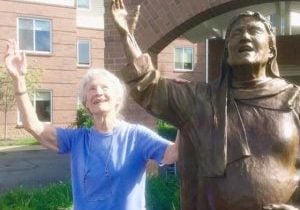 A white-haired woman raises her hand and looks to the sky with joy in an identical copy of the pose of the statue next to her.