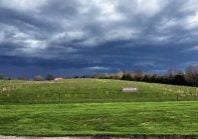 The Loretto Motherhouse entrance in Kentucky. A sprawling bright green grassy hill with a fence to keep in the cows and a sign that says "Loretto Motherhouse" in the distance. Half the frame are large pillowy clouds on a darker blue and grey day.