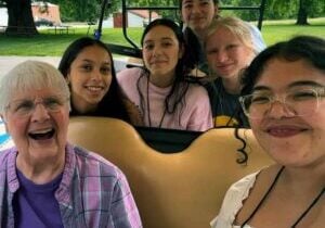 A group of five high school girls smile with a nun on a golf cart outside.