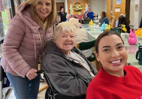 Three women smiling for a photo. One woman stands behind an older woman in a wheelchair and another woman sits at the communal table.