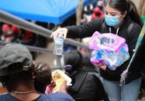 A young Latina in a mask reaches up the stairs to hand a bottle of water to a Black child eating on her caregivers lap.