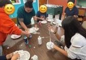 A volunteer at a table with a bunch of elementary aged kids shown making slime together.