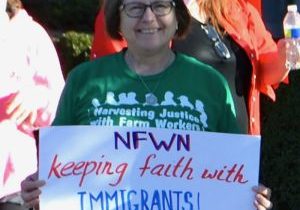 A woman wearing a green shirt and a smile stands amongst a crowd and holds a sign that reads "NFWN keeping faith with Immigrants", with the National Farm Worker Ministry logo in the lower right corner.