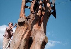 A woman stands on a ladder to work near the top of a huge sculpture. The base is shaped like a tremendous tree trunk, and a bird with black metal wings hovers over it.