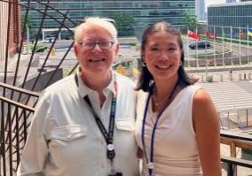 A middle aged woman with white hair, black glasses and a white polo smiles with a college aged women with shoulder length brown hair a white tank top and silver hoop earrings. They are in front of the United Nation. Behind them you can see an array of various nations flags.