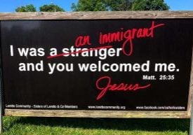 Large black sign with "I was a stranger and you welcomed me. Matt. 25:35" in white text, with "a stranger" crossed out with red and "an immigrant" written over it in red. "Jesus" is signed in red.