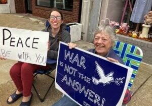 An elder nun sitting in a lawn chair on the corner of an intersection sits next to a younger woman and they are both holding signs of peace. One says "Peace be with you" the other says "War is not the answer"