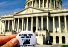 A hand holds a button that reads "Work for justice; act for peace." A capitol building is visible in the background.