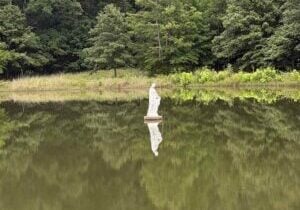 A statue of the Virgin Mary stands in the middle of a lake with the reflection of the woods behind her.
