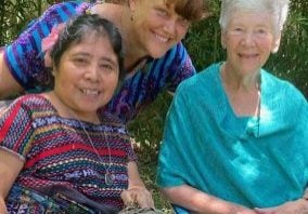 Three women, one in traditional Guatemalan patterns, lean in for a photo together in the dappled shade.