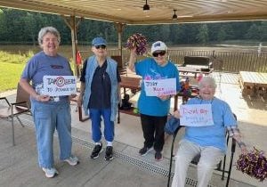 Four elder women standing and sitting under an awning holding posters on a 5k race for the runners. The posters say "Dont stop people are watching" "Just keep running" and "tap here to power up"