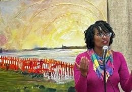 A black woman with natural hair, a pink shirt and glasses stands in front of her mixed media piece she made during an artist residency. The piece has green grass, a red fence, an ocean and a bright yellow sky.