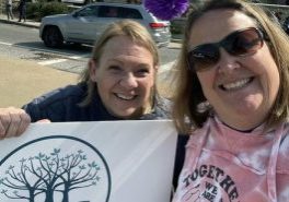 Two women, one wearing sunglasses and one holding a large sign with a tree design, smile for a selfie together outdoors on a sunny day.