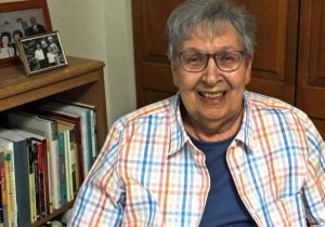 An older white woman sitting down smiling wearing glasses a blue t-shirt and a striped blue and orange collared long sleeve shirt over the blue shirt. She looks relaxed and is sitting next to her books on a bookshelf and on top of the shelf are family photos.