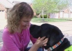 A woman in a pink shirt with light brown shoulder-length wavy hair is nose to nose with her seeing-eye dog as she holds his face in her hands.