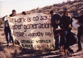 Photo from 1989 of eight people holding up a large fabric banner amongst the Nevada scrub desert. The banner reads "Our work is to sow. Another generation will reap. - Dorothy Day Catholic Worker, Denver, Colorado