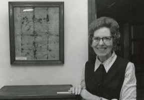 Archival black and white photo of a woman in glasses standing next to a framed historical document.