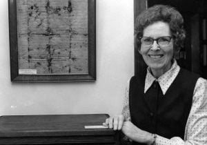Archival black and white photo of a woman in glasses standing next to a framed historical document.