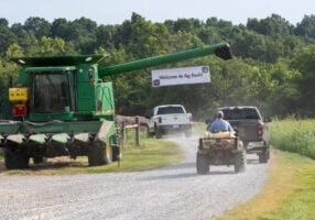 A sign hanging on a tractor that says "Welcome to Ag Bash" that is hanging over a dirt road. Two pickup trucks and a man on a small tractor drive on the road to the Ag Bash.