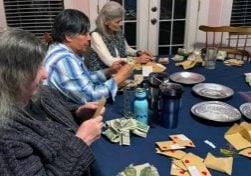 Three older women, two with long grey hair and one with shorter black hair are sitting at a table counting donations collected by students. The donation envelopes are decorated with hearts.
