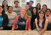 A large group 16 Latino college students of varying ages and one white woman in her 40s smiling together at an end of year retreat.