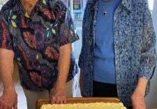 Two women in colorful blouses and short white hair and glasses smile together in front of a yellow cake that says "Happy 90th Birthday Joan and Ellen!"