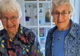 Two women in colorful blouses and short white hair and glasses smile together in front of a yellow cake that says "Happy 90th Birthday Joan and Ellen!"