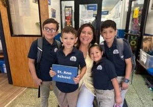 Four elementary school students with blue polo shirts and khakis taking a picture with a teacher who is squatted down to be the student's height. One of the students is carrying a blue plaque that says "St. Ann School"
