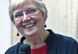 Short haired woman wearing glasses, a red shirt and navy blue jacket smiles for a picture while using a microphone.