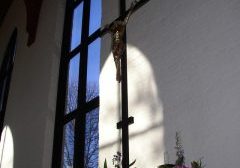 Light from an arched window illuminates a tall crucifix in a church. Spring flower arrangements surround the base of the cross.