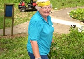 Woman with short, white curly hair and a yellow bandana pauses while weeding to smile for a photo.