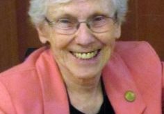 A woman with short white hair and wire glasses smiling brightly for a headshot picture wearing a black blouse and salmon pink blazer in front of a plain dark brown wall.