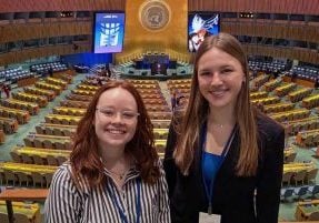 Two young college aged students smilng together in front of the UN general assembly hall. Behind them is an impressive room with hundreds of seats and the UN emblem centered over the podium with two large screens on each side.
