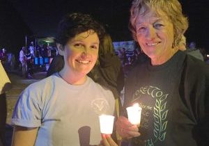 Two women stand outside in the night, holding votive candles.