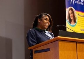 A young Black woman smiling brightly as she stands and talks at a podium. She is wearing a blue sweatshirt and a silver necklace with her hair brown shoulder length hair straightened and pulled back with a clip on one side.