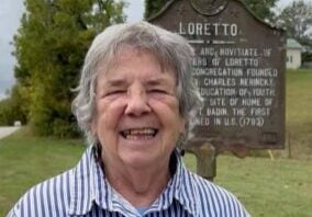 An older woman smiles for the camera while standing outside next to a sign titled "Loretto" followed by details of the Sisters of Loretto history.