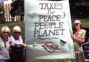 Six women outdoors on a sunny day wearing sunglasses and hats while standing next to and holding a large poster stating: Taxes for Peace People Planet.