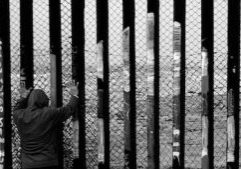 A black and white photo of somebody in a hoodie, shorts and sneakers back to the camera, putting their hands on the border wall between the U.S. and Mexico.