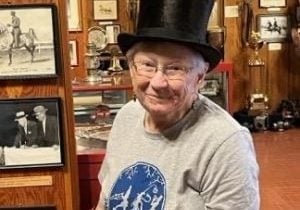 Older woman wearing a "Music Folk" t-shirt and top hat stands in a display of old photographs.