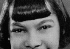 A black and white headshot picture of a young woman with straight black hair with bangs. She is smiling brightly in front of a plain background.
