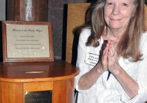 A woman with her hands clasped in gratitude in front of her stands next to a donation box in a chapel.