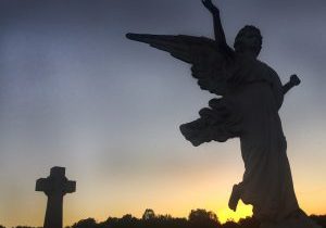 An angel and a cross are profiled in front of the setting sun.