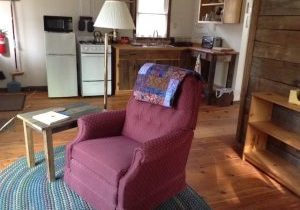 A red chair with a colorful quilt on top sits centered in the room of a small wood cabin.