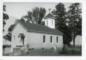 Black and white archival photo of a white wooden church with a bell tower and dark roof. Headstones can be seen in the foreground.