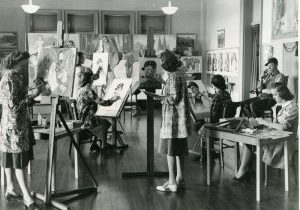 Students in an art class, standing at easels and painting portraits of a man smoking a pipe.