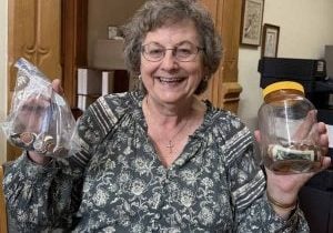 A woman with a huge smile stands holding a bag of coins in her right hand and a jar of coins and bills in her left.